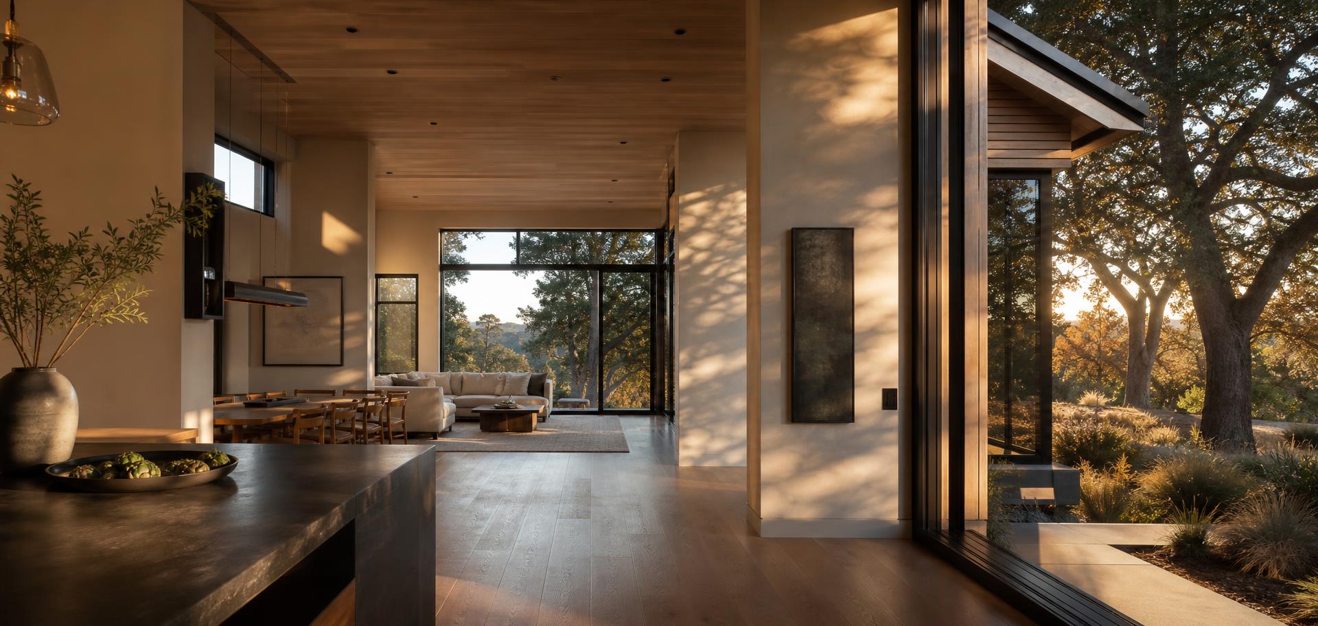 Open-plan living space in a modern Northern California home with oak floors, floor-to-ceiling black steel windows looking onto oak trees, and late afternoon sunlight raking across plaster walls